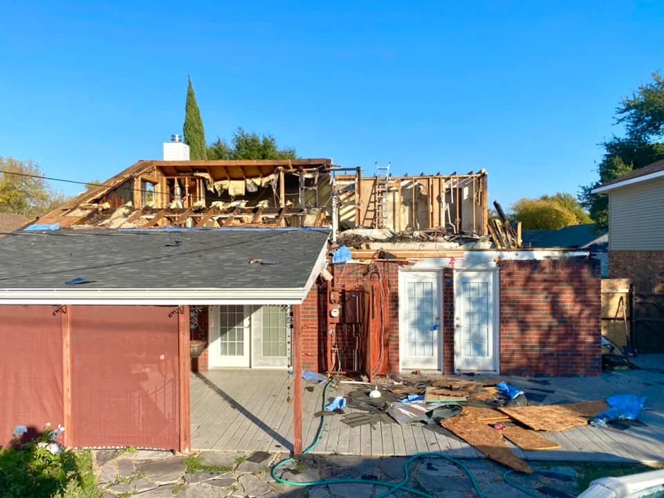 House undergoing renovation; roof partially removed, exposing wood framing, brick and concrete patio below.