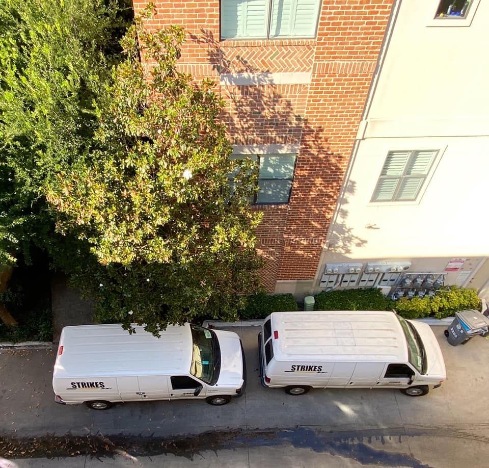 Two white vans parked near a brick building with windows and a tree. One van says 