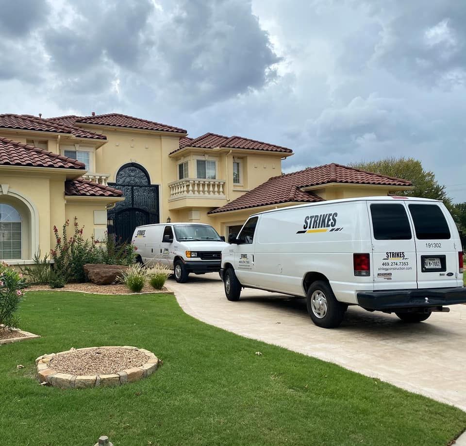 Two white vans parked in front of a large, yellow house with a red-tiled roof. Overcast sky.