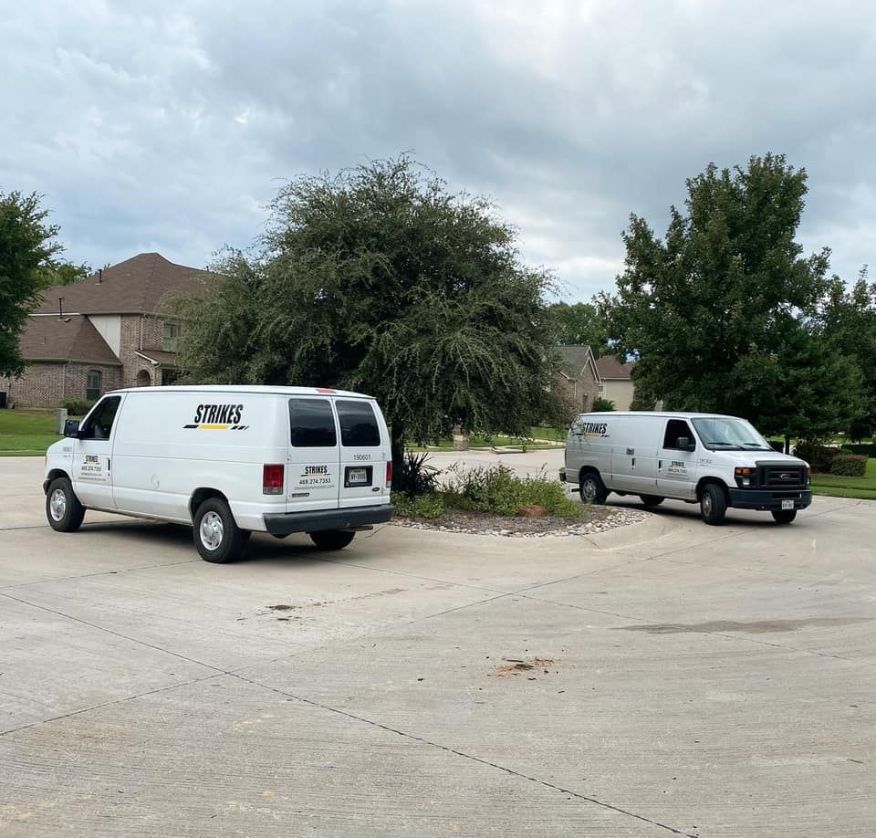 Two white service vans parked on a concrete lot, with the same logo. A house and trees are in the background.