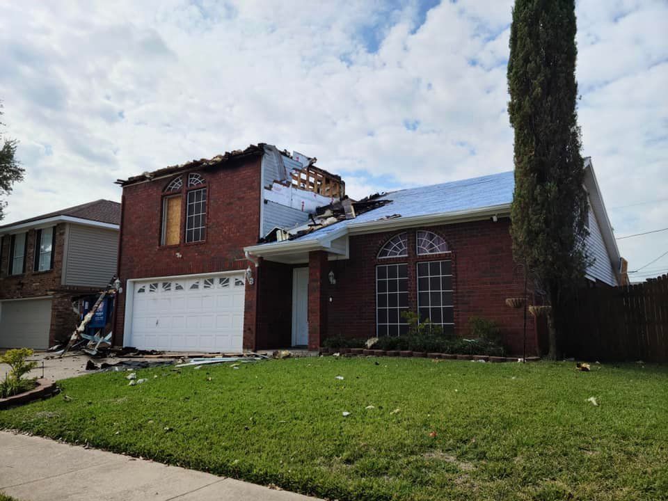 House with significant roof damage, debris scattered on the lawn, likely from a storm.