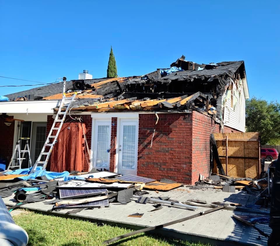 House with fire damage; charred roof, brick exterior, debris on the ground, ladder leaning against the structure.