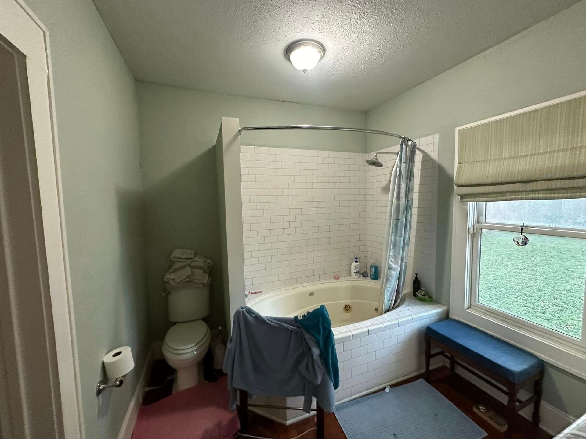 Bathroom with a tub/shower, toilet, and window; green walls, white tiles, and a blue bench.