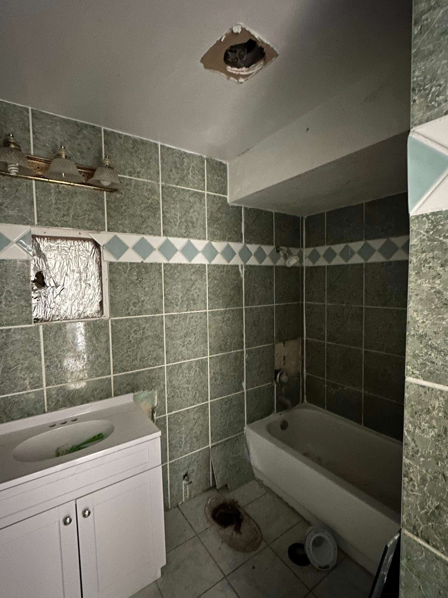 Bathroom with green tile, white vanity, and a tub. Damaged ceiling and a small window.