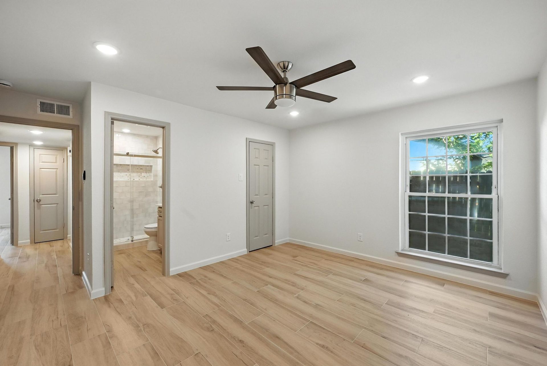 Empty bedroom with wood-look floors, white walls, ceiling fan, and window. Bathroom doorway visible.