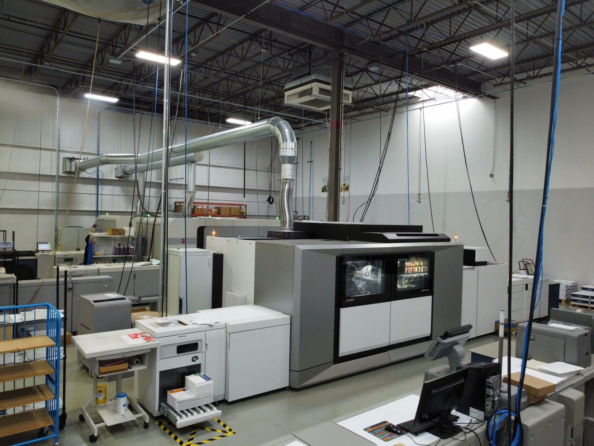 Large printing press in a warehouse setting, with ventilation system overhead and various support equipment nearby.