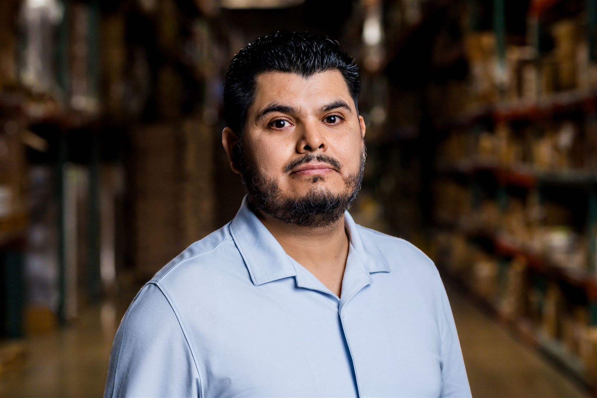 Man standing in a store aisle, wearing a light blue shirt, with shelves blurred in the background
