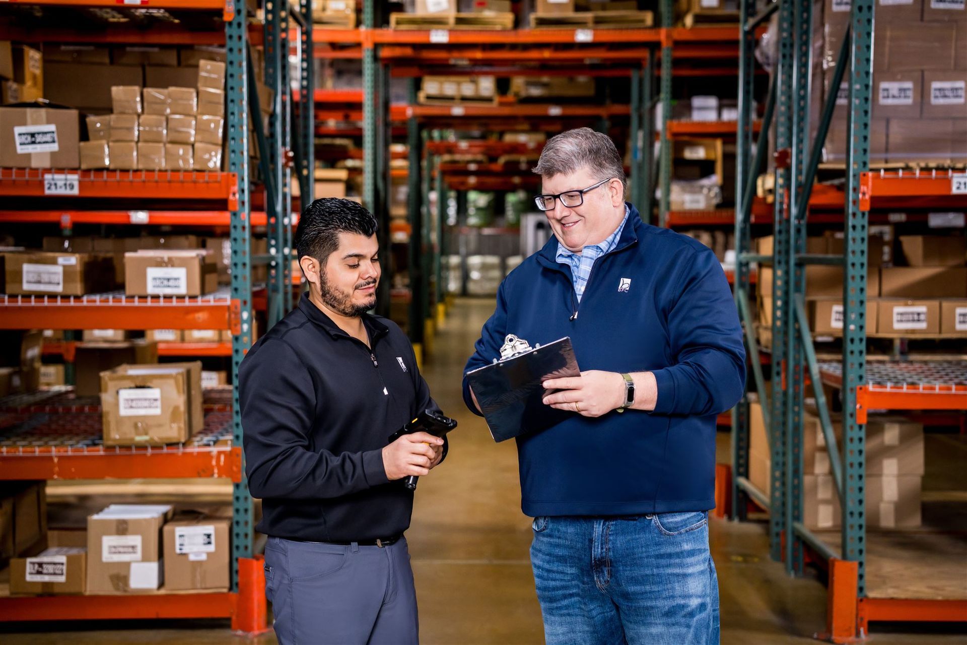 Two workers in a warehouse discuss a clipboard in front of stocked shelves.