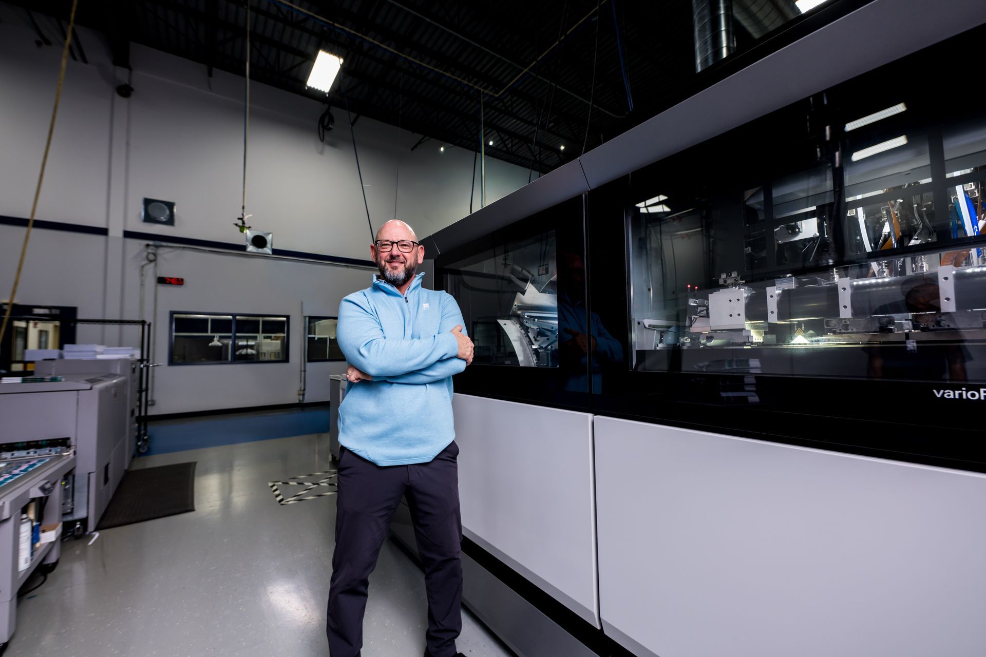 A person stands with arms crossed in front of a large, industrial printing machine inside a bright, spacious facility.