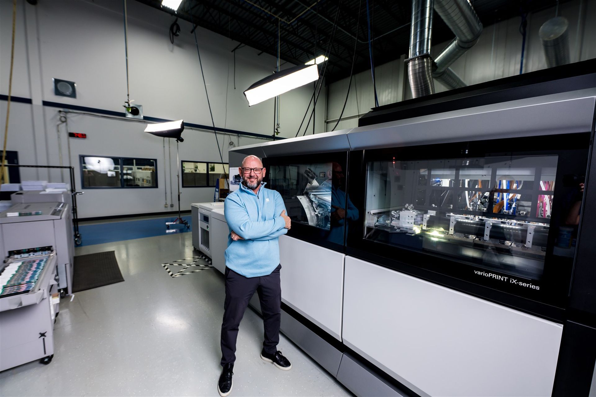 A person stands with arms crossed in front of a large, industrial printing machine inside a bright, spacious facility.