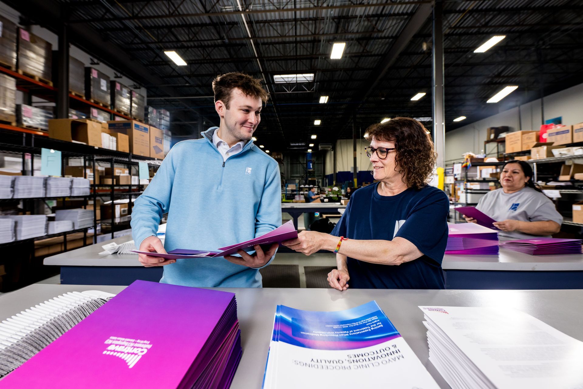 Two workers review printed materials in a warehouse, with colorful stacks of brochures on the table.