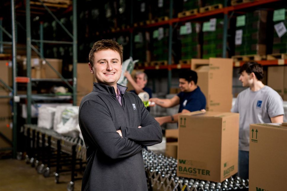 Warehouse worker smiling with arms crossed beside stacked boxes and bottles on a production line