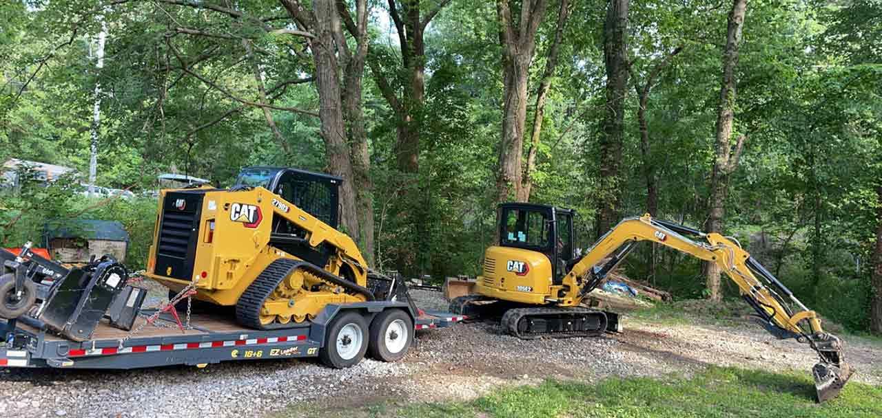 Two excavators are on a trailer in the woods.