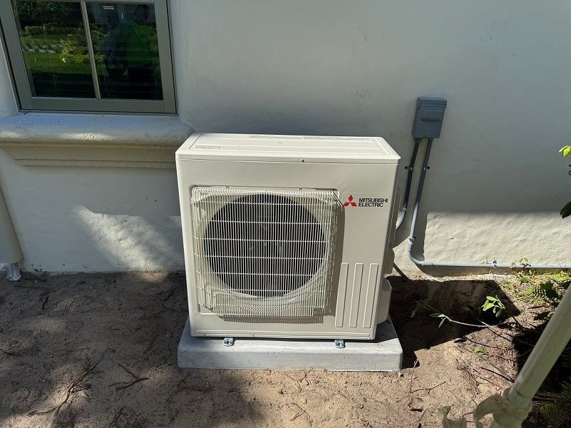 Mitsubishi outdoor AC unit on a concrete pad next to a light-colored wall and window.