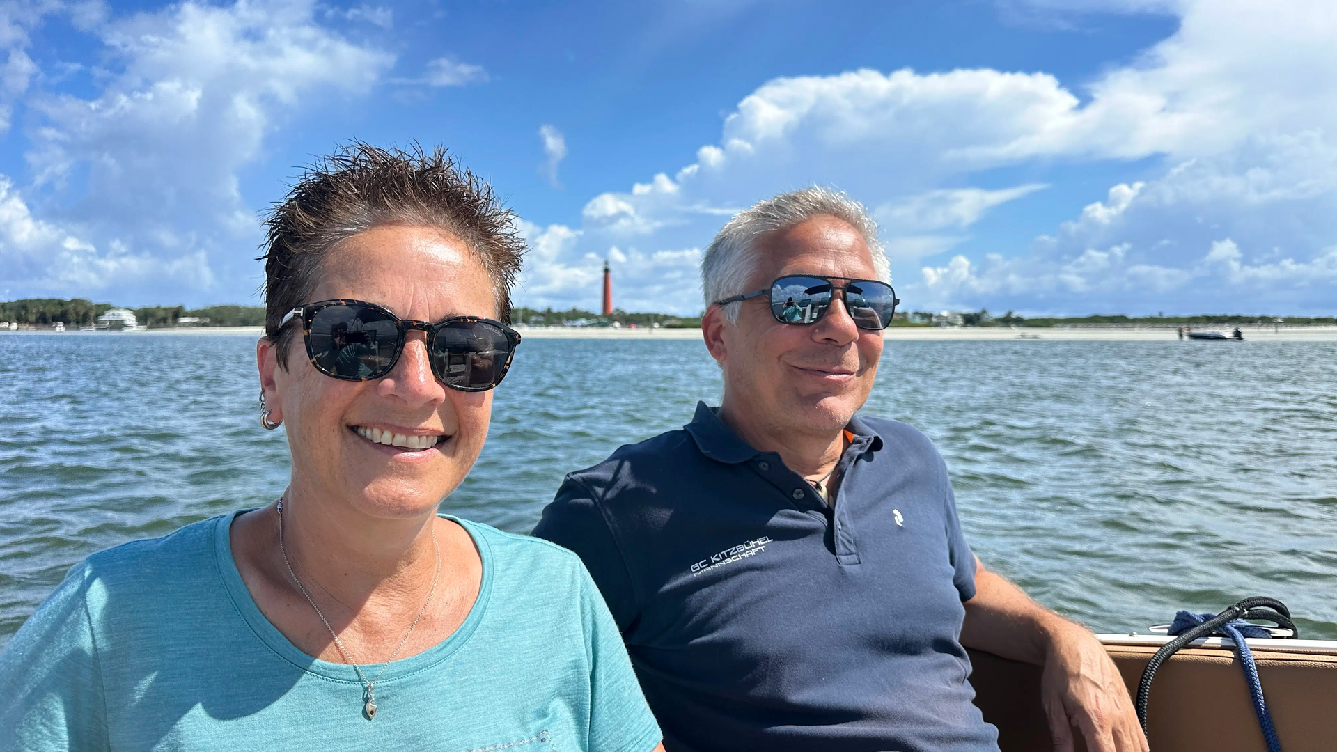 Two people in sunglasses smiling on a boat on a sunny day, with a lighthouse in the background.