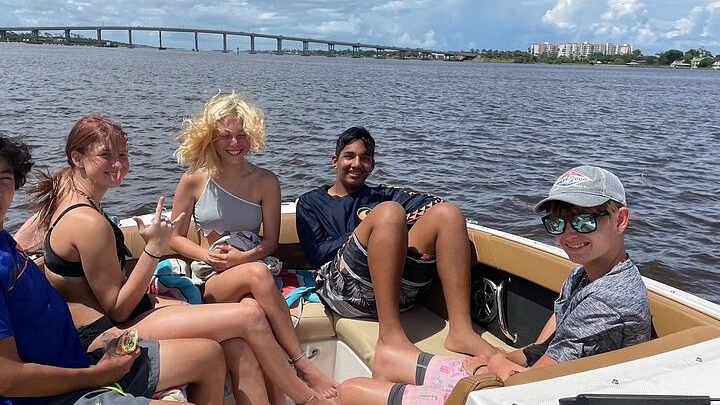 Group of teens on a boat, smiling and relaxing on the water. A bridge is in the background.