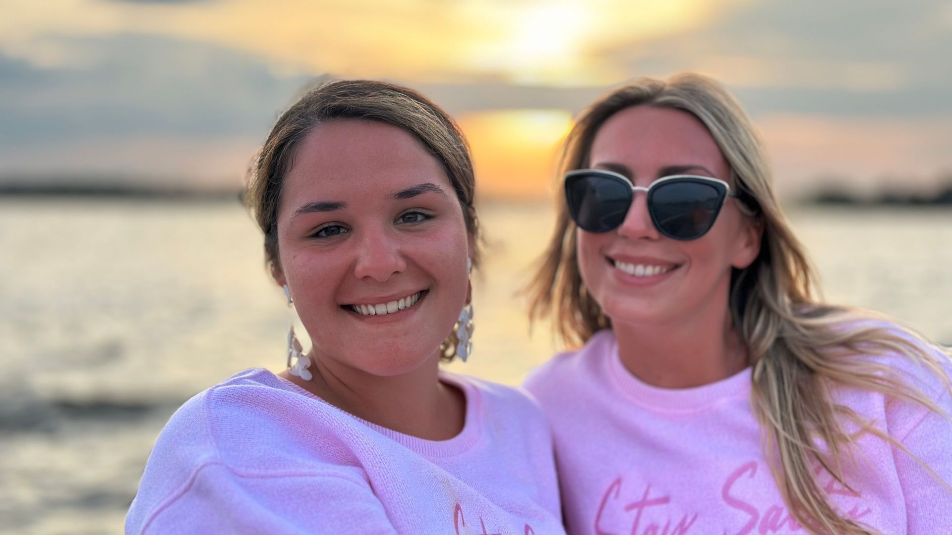 Two smiling women in pink sweatshirts pose outdoors at sunset. One wears sunglasses, and the other has large earrings.