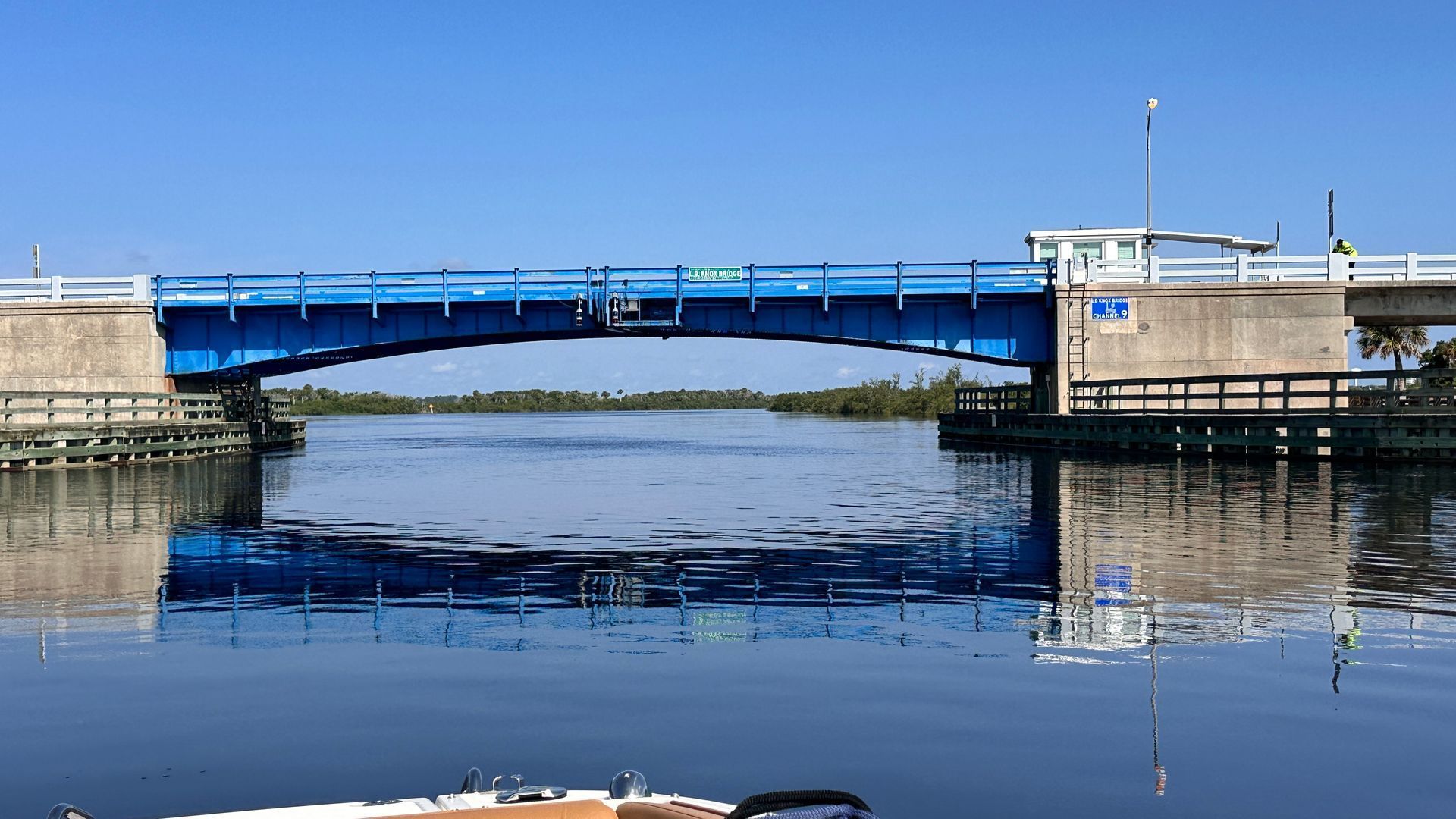 Blue drawbridge over a calm waterway, reflecting in the water. Bright blue sky above.