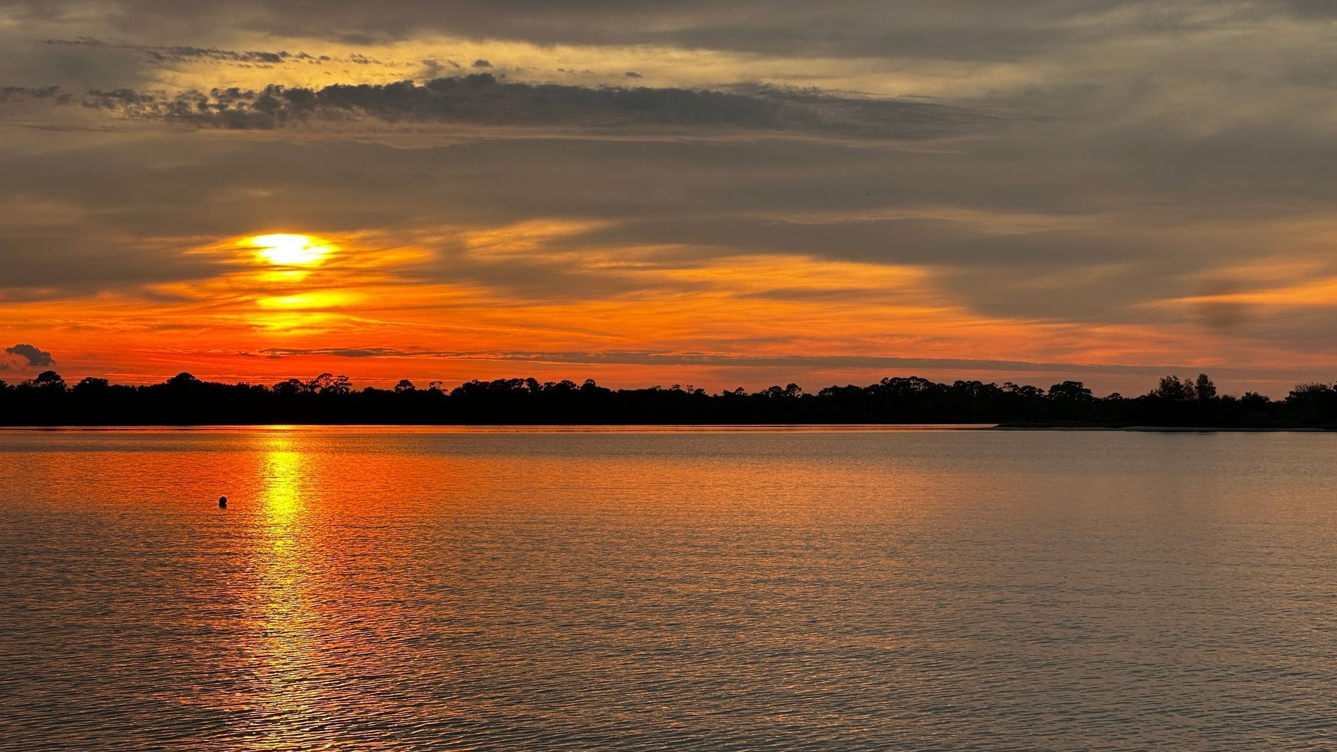 Sunset over calm water, reflecting orange and yellow light. A dark treeline is visible in the distance.