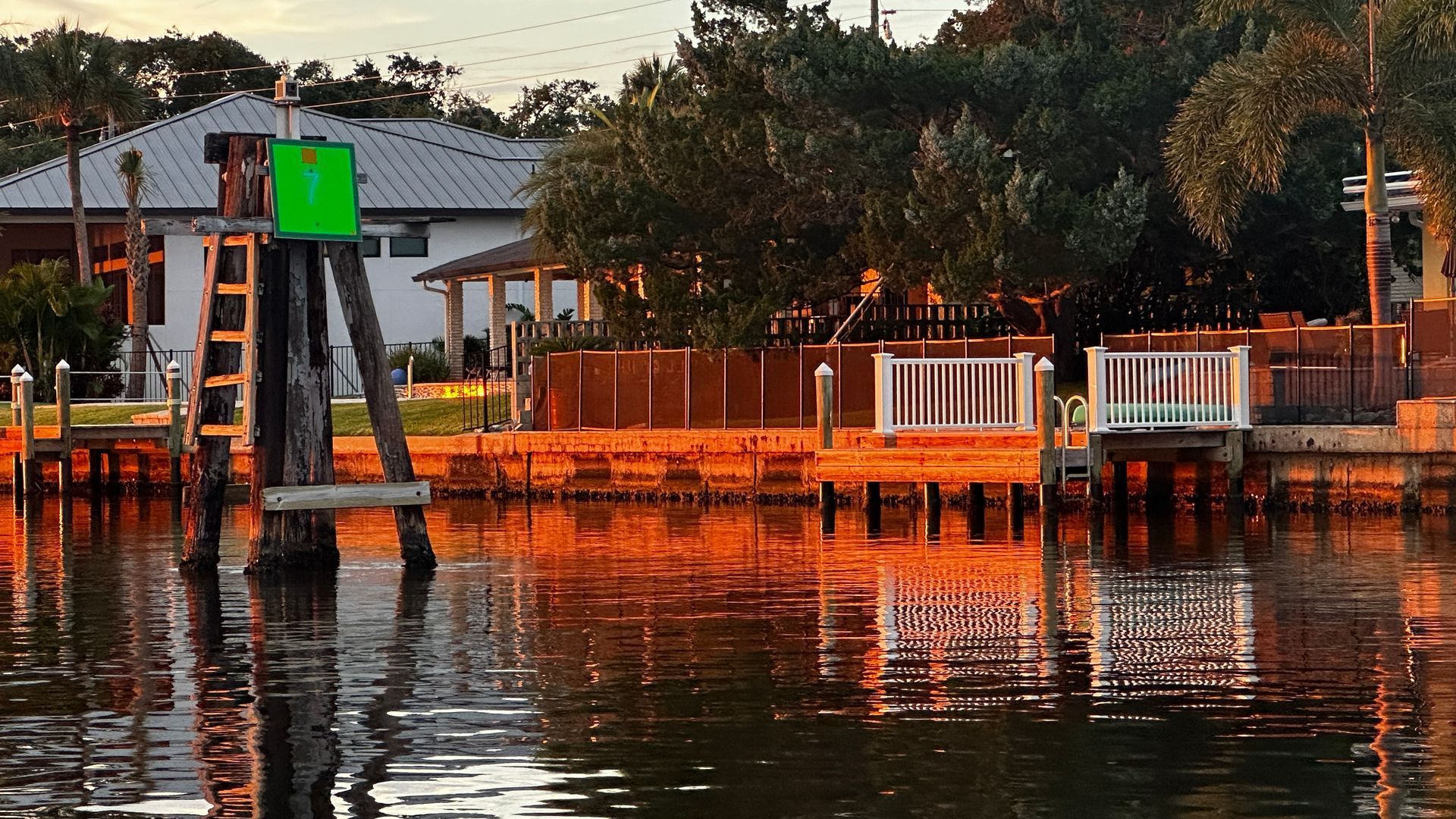 Waterfront scene with a green channel marker and docks reflecting golden sunset hues, with a house in the background.