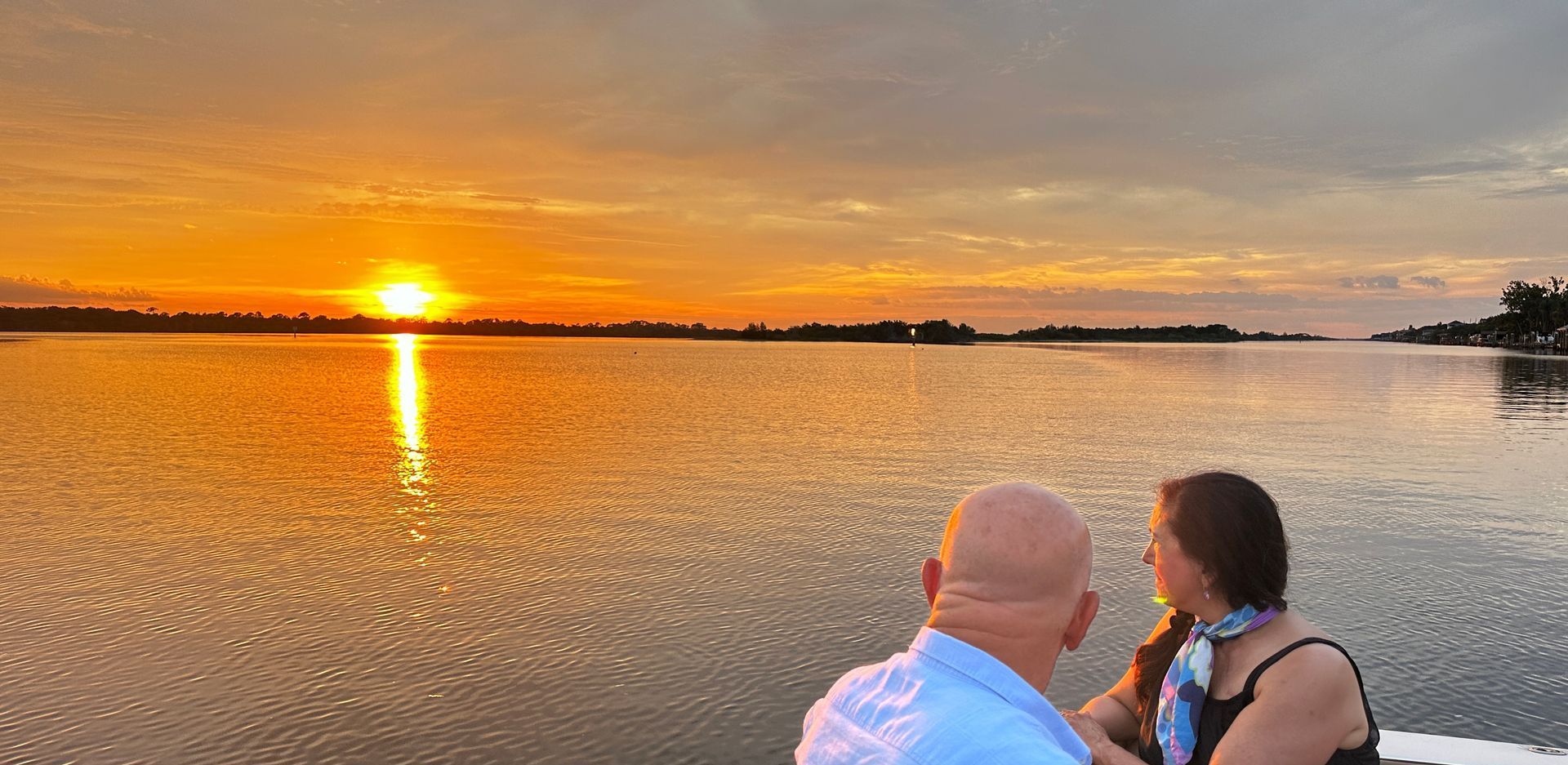 A couple watches a vibrant sunset over calm water. The golden sun reflects on the water.