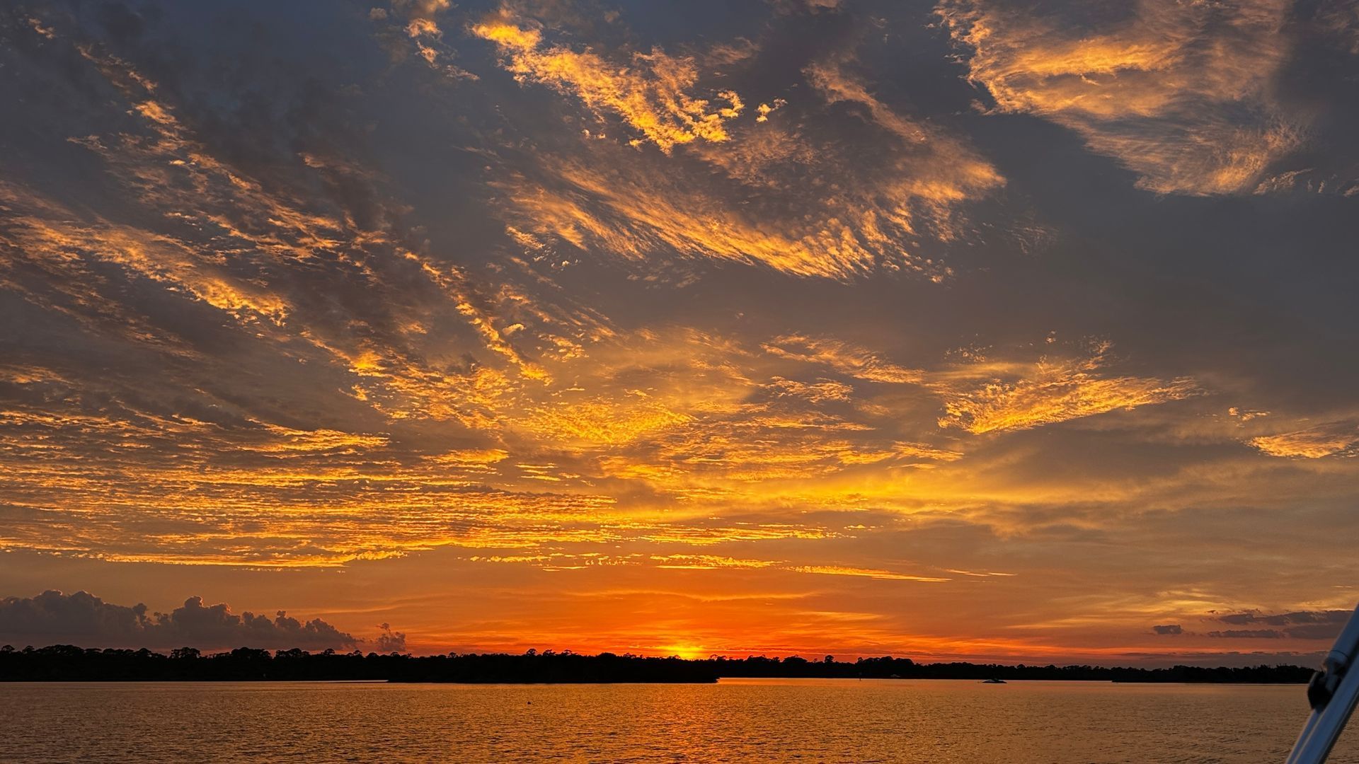 Sunset over water, with a vibrant orange and yellow sky dotted with clouds; dark horizon silhouette.