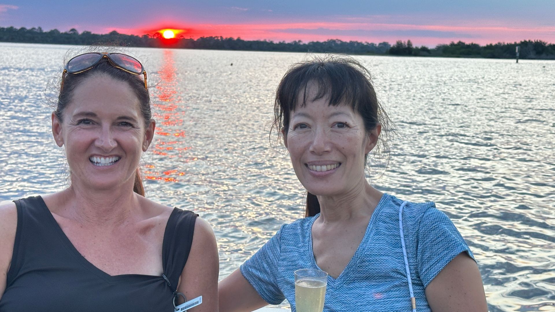 Two smiling women pose in front of a sunset over water. One holds a champagne flute.