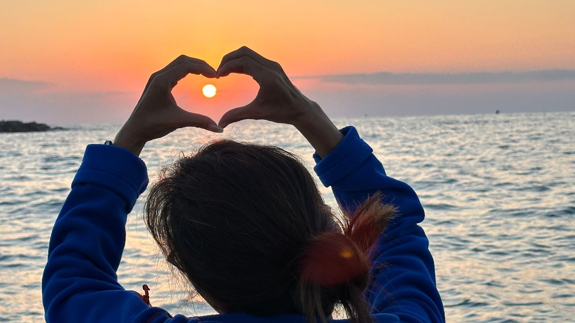 Person making a heart shape with hands, framing the setting sun over the ocean at dusk.