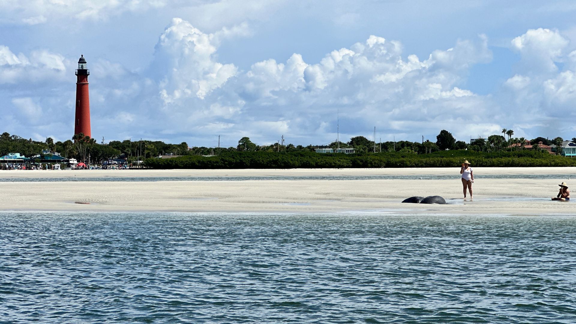 A beach scene with a red lighthouse, blue water, and people on the sand under a cloudy sky.