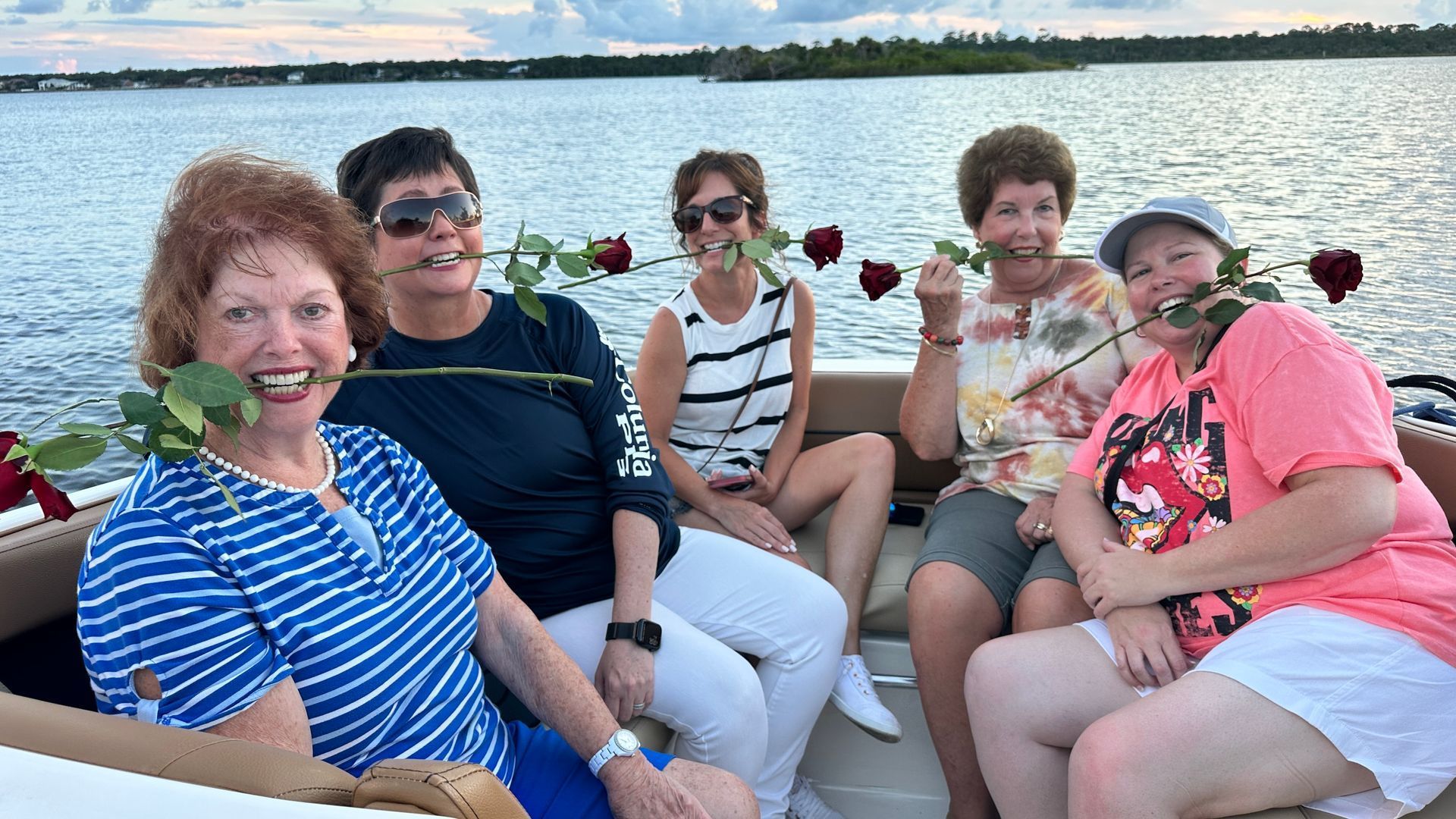 Five women on a boat holding red roses in their mouths, smiling at the camera. 