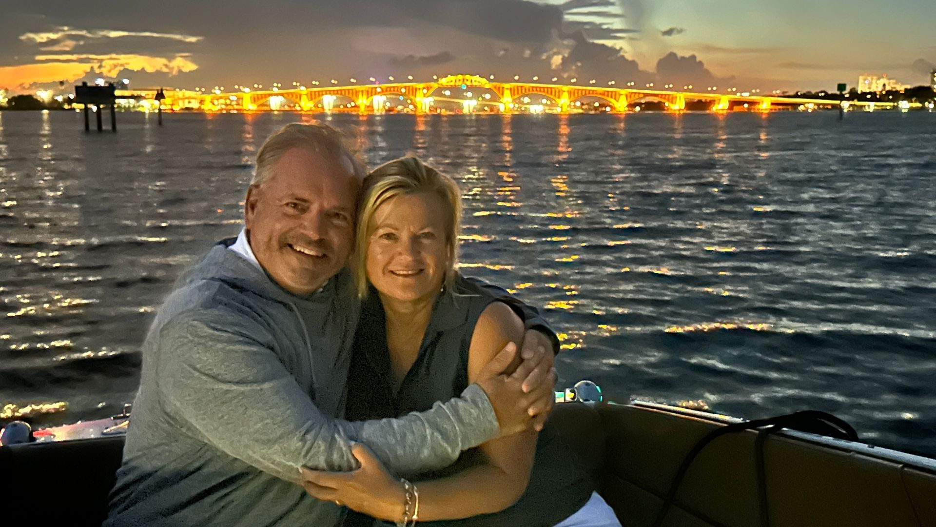 Smiling couple embracing on a boat at dusk, with a brightly lit bridge and water in the background.