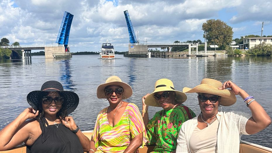 Four women in sun hats and sunglasses smile on a boat, with a blue drawbridge in the background.