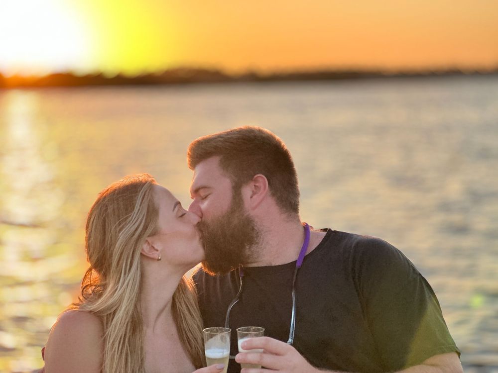 Couple kissing on a boat on a waterway with houses in the background. The woman has orange braids, the man wears a hat.