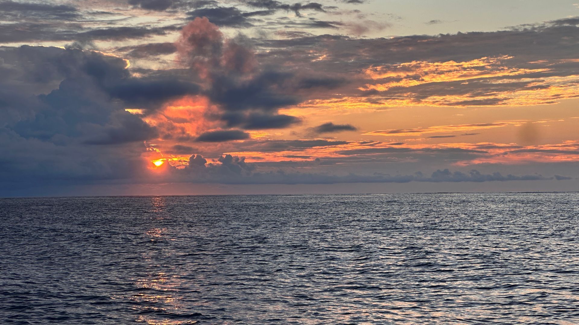 Sunset over ocean; sun peeks through clouds, casting gold and orange hues on the water.