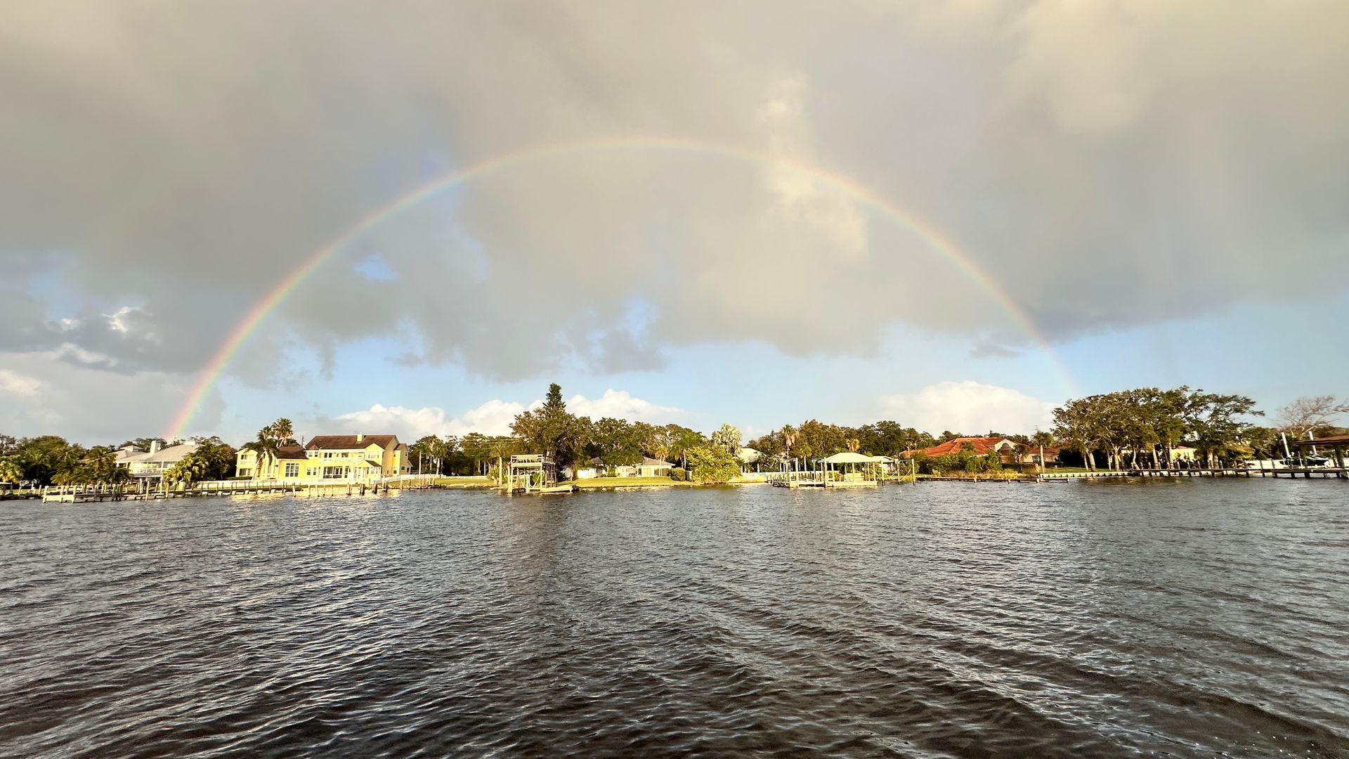 Rainbow arcing over a lake and shoreline of houses and trees under a cloudy sky.