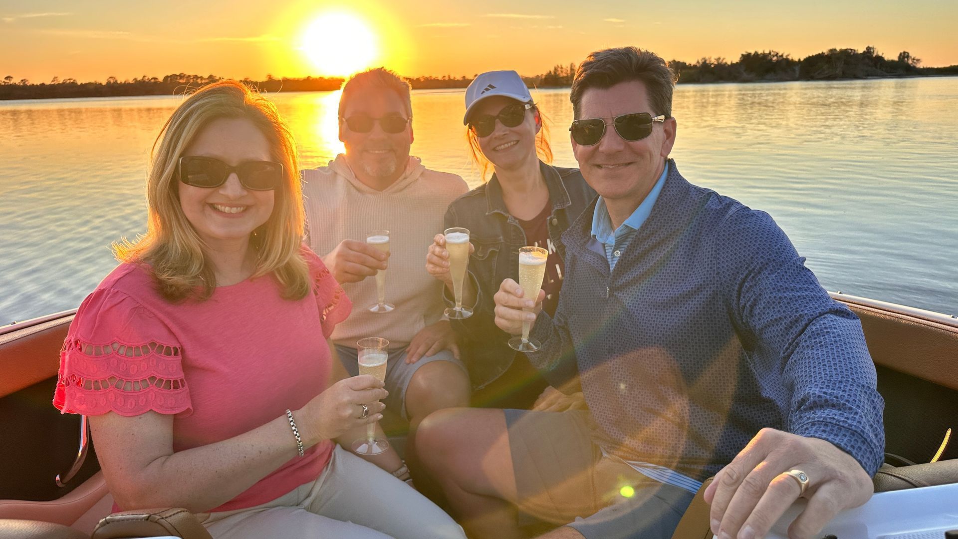 Four people on a boat, toasting with champagne at sunset. They're smiling, enjoying the water and golden light.
