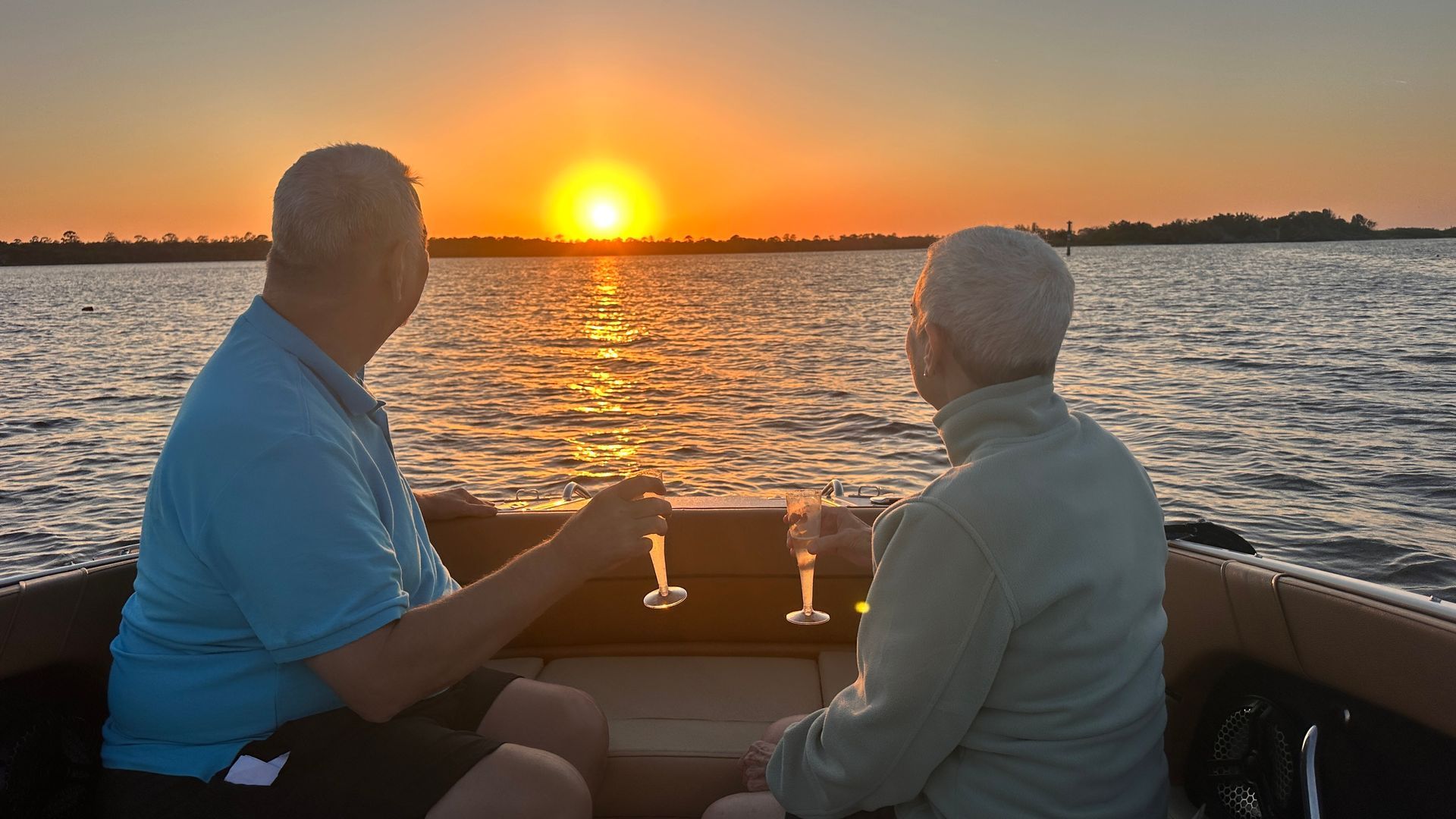 Two men on a boat at sunset, toasting with glasses. The sun sets over the water, casting orange and gold reflections.
