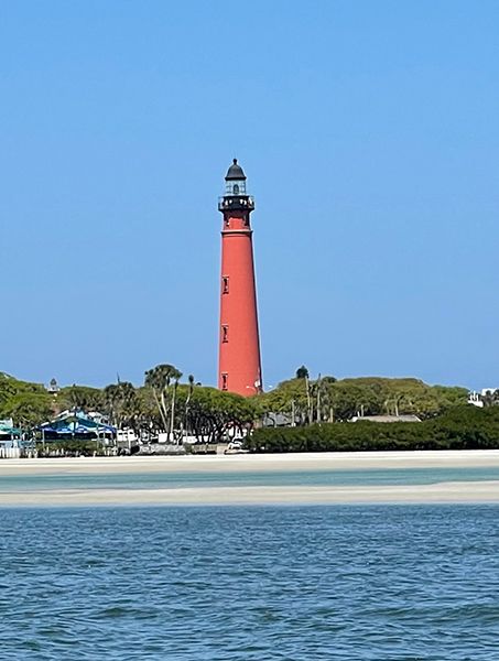 Red lighthouse on a beach under a clear blue sky. Trees and buildings line the shore near the water.