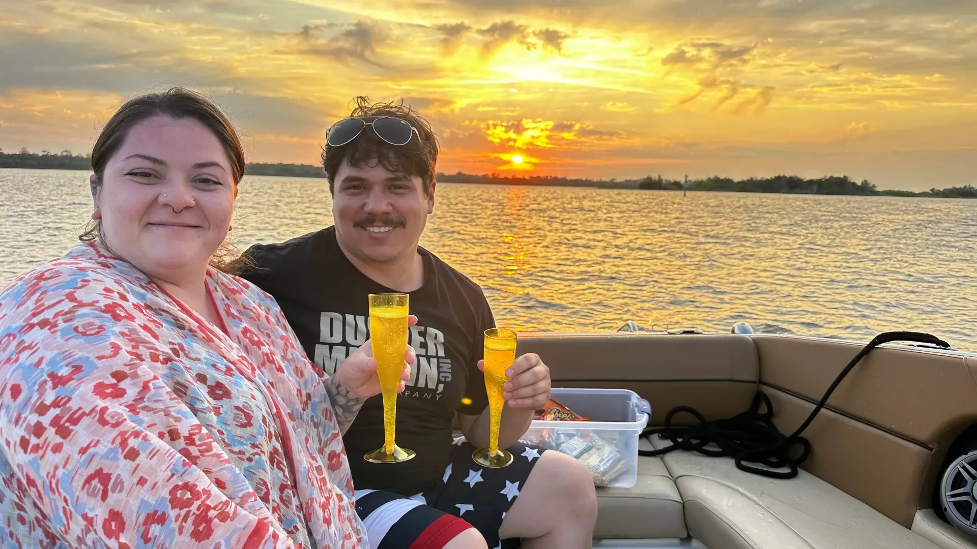 Couple on a boat at sunset, toasting with champagne. The sun is setting over the water, creating a golden glow.