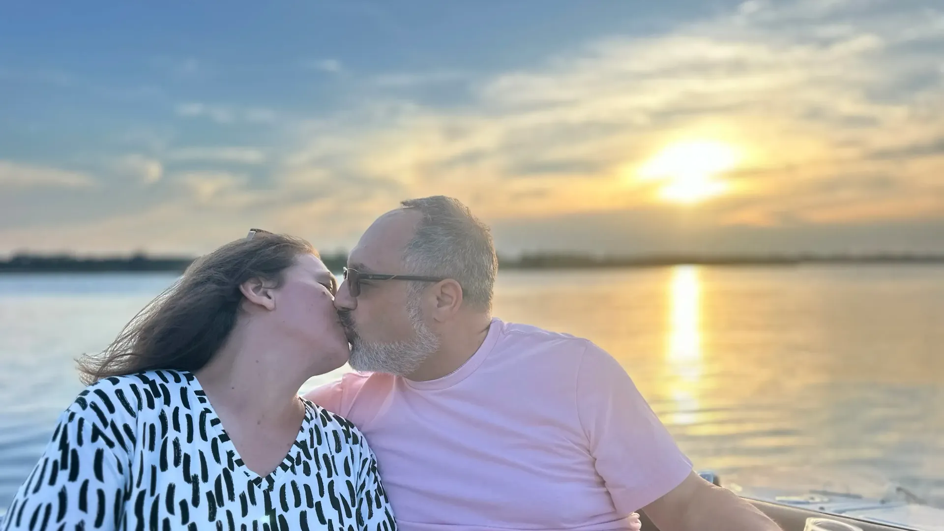 Couple kissing at sunset over a body of water; man in pink shirt, woman in black and white top.
