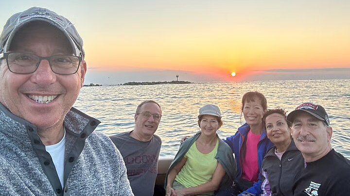 A group of people smiling on a boat at sunset. The sun is a bright orange ball over the water.