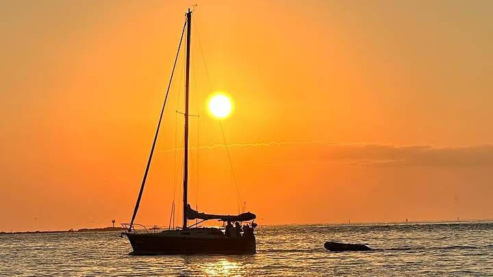 Sailboat silhouetted against an orange sunset on the water. The sun is centered above the boat's mast.