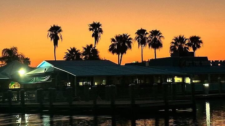 Silhouette of buildings and palm trees at sunset over a body of water; orange and yellow sky.