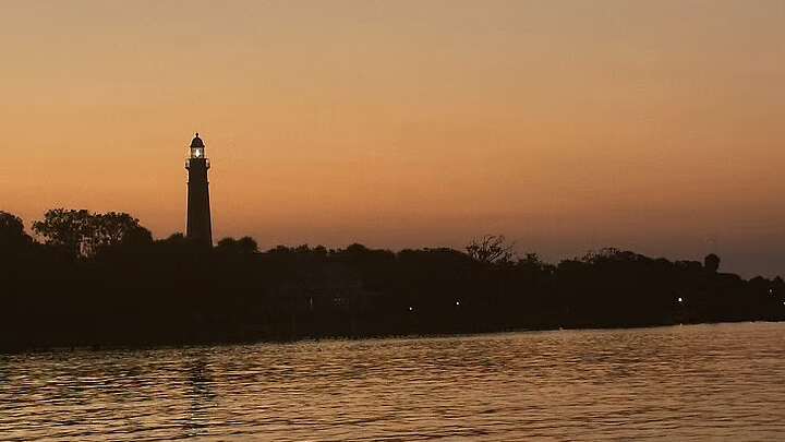 Silhouette of a tall tower with a light atop against an orange and yellow sunset sky, reflected in the water.