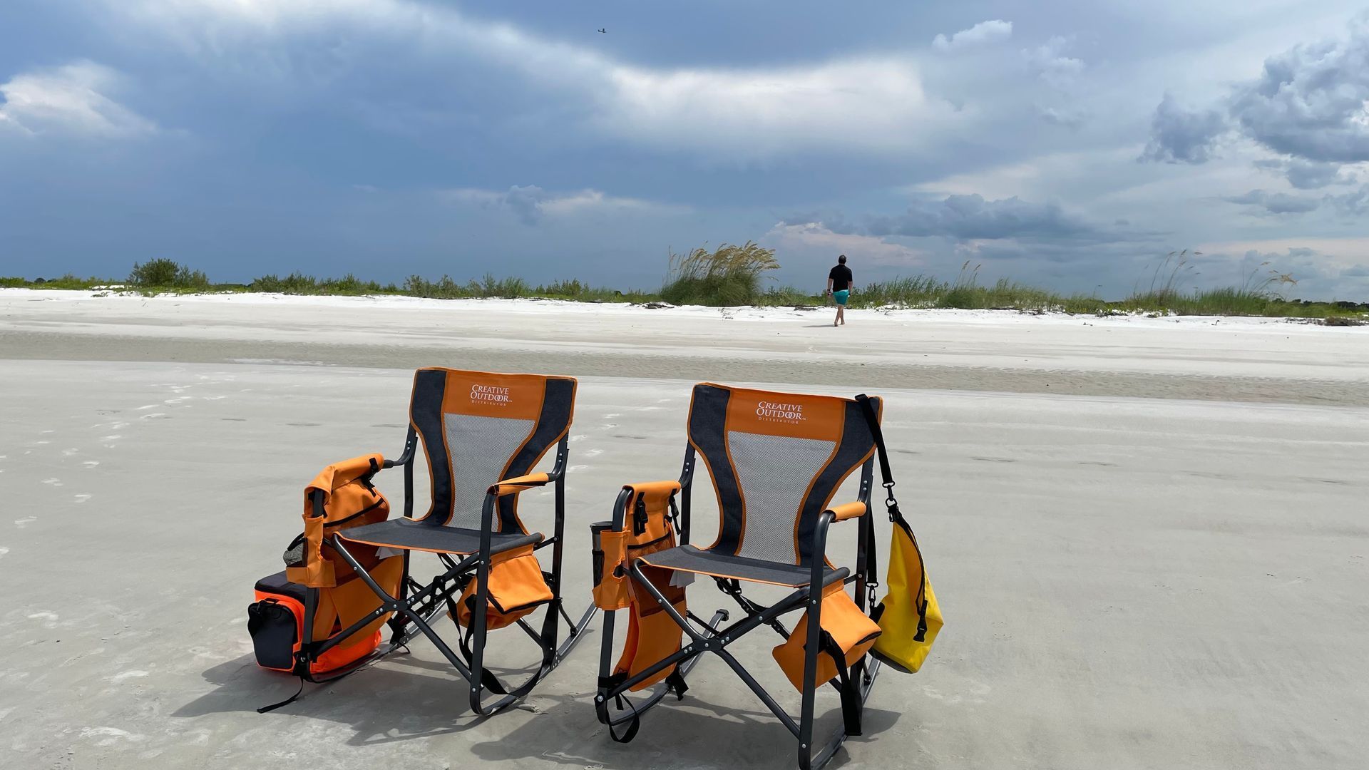 Two orange and black beach chairs on a white sandy beach with a person walking in the distance under a cloudy sky.