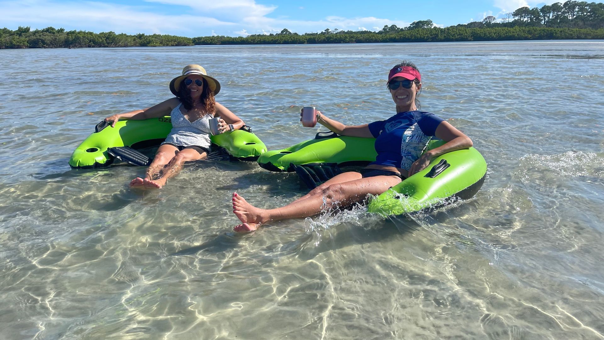 Two women floating on green inner tubes in clear water, enjoying a sunny day. One wears a hat, the other a visor.