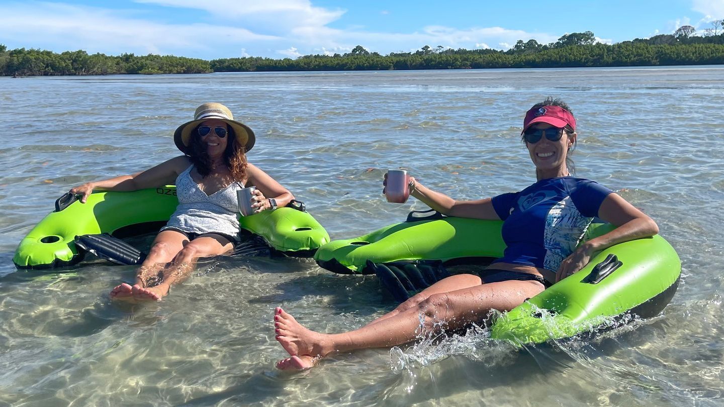 Two women relax in neon green inflatable tubes in clear, shallow water; one holds a mug.