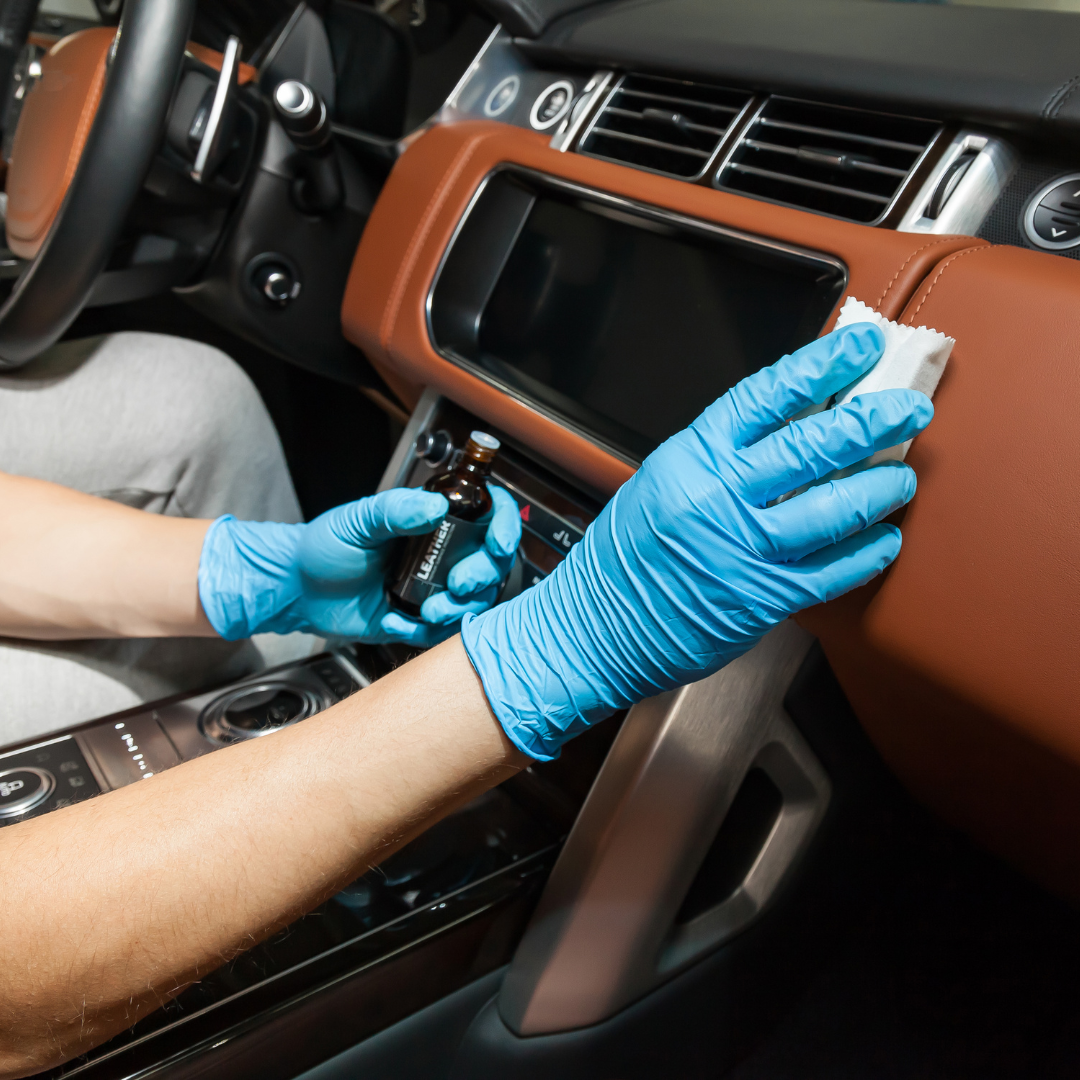 A person wearing blue gloves cleans the brown leather dashboard of a car with a wipe and bottle.