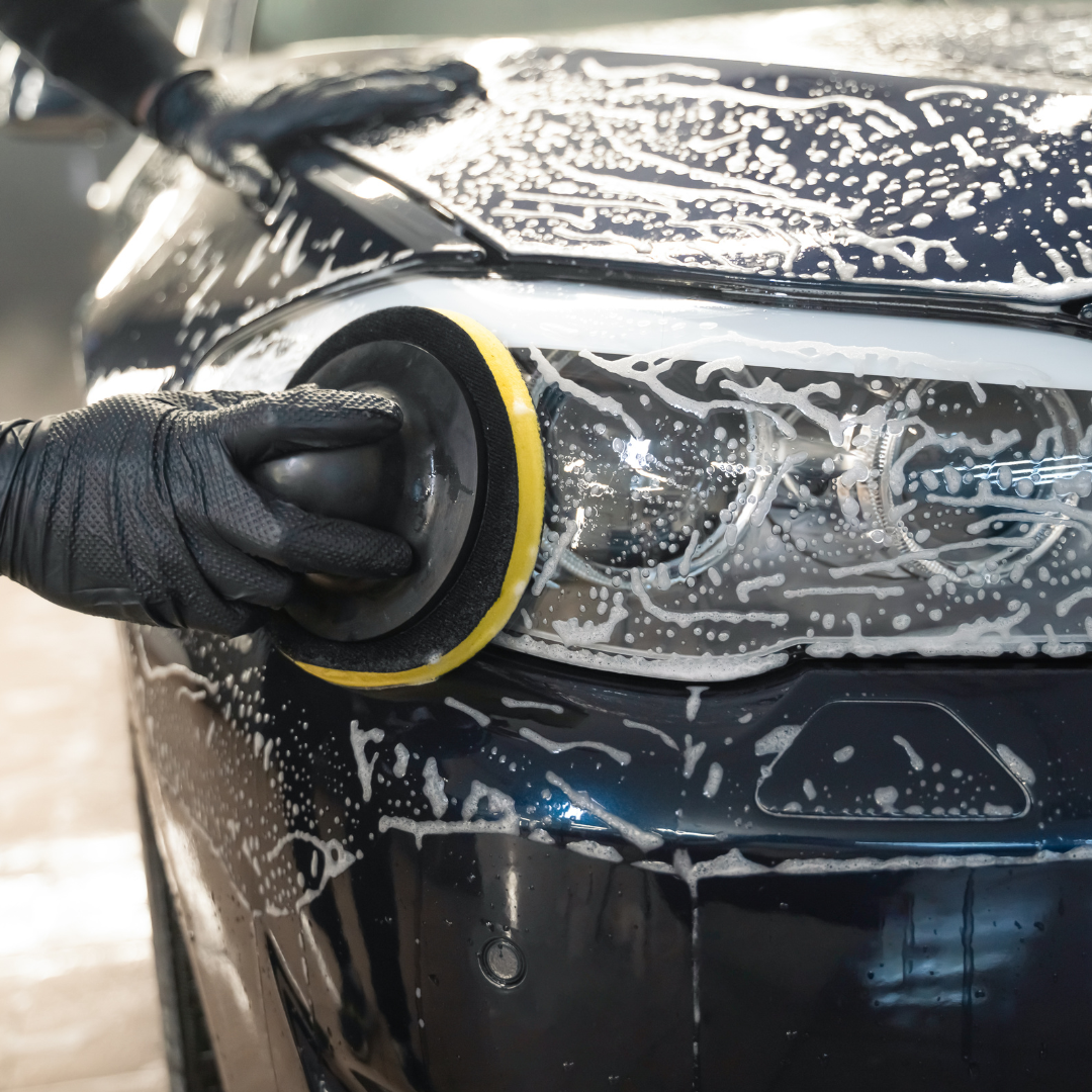 Person polishing a dark car's headlight with a machine, covered in soapy foam and wearing black gloves.