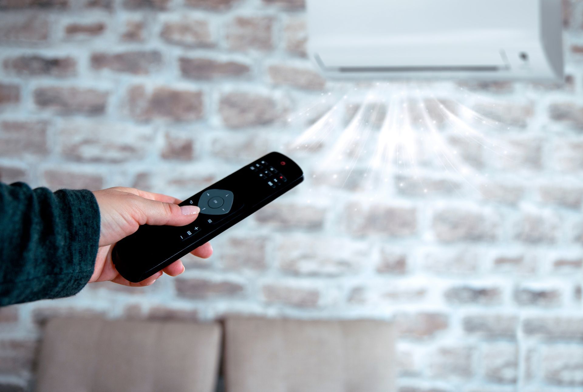 Hand holding a remote control pointing towards an air conditioner on a brick wall.